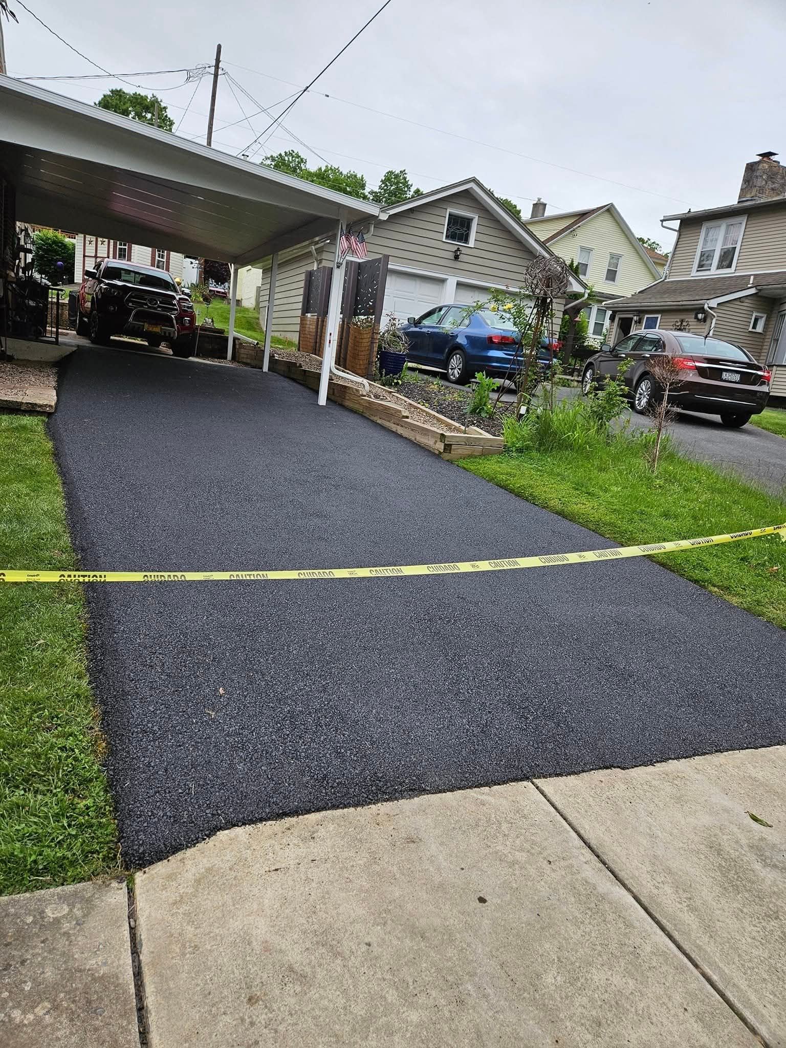 A newly paved dark gravel driveway with a covered parking area, blocked off by yellow caution tape in a residential area.