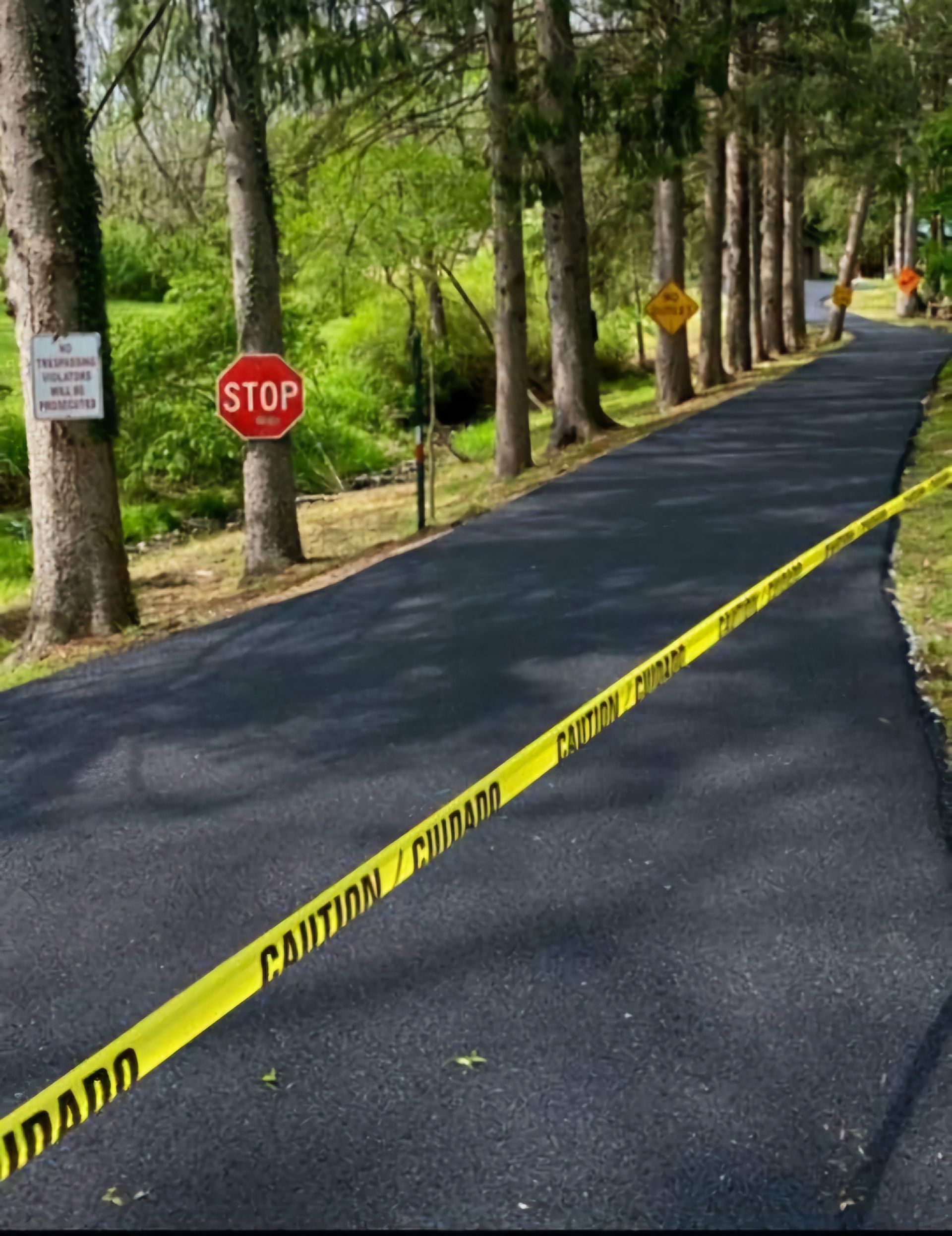 A freshly paved road is blocked by yellow caution tape, with a red stop sign and other warning signs nearby in a wooded area.