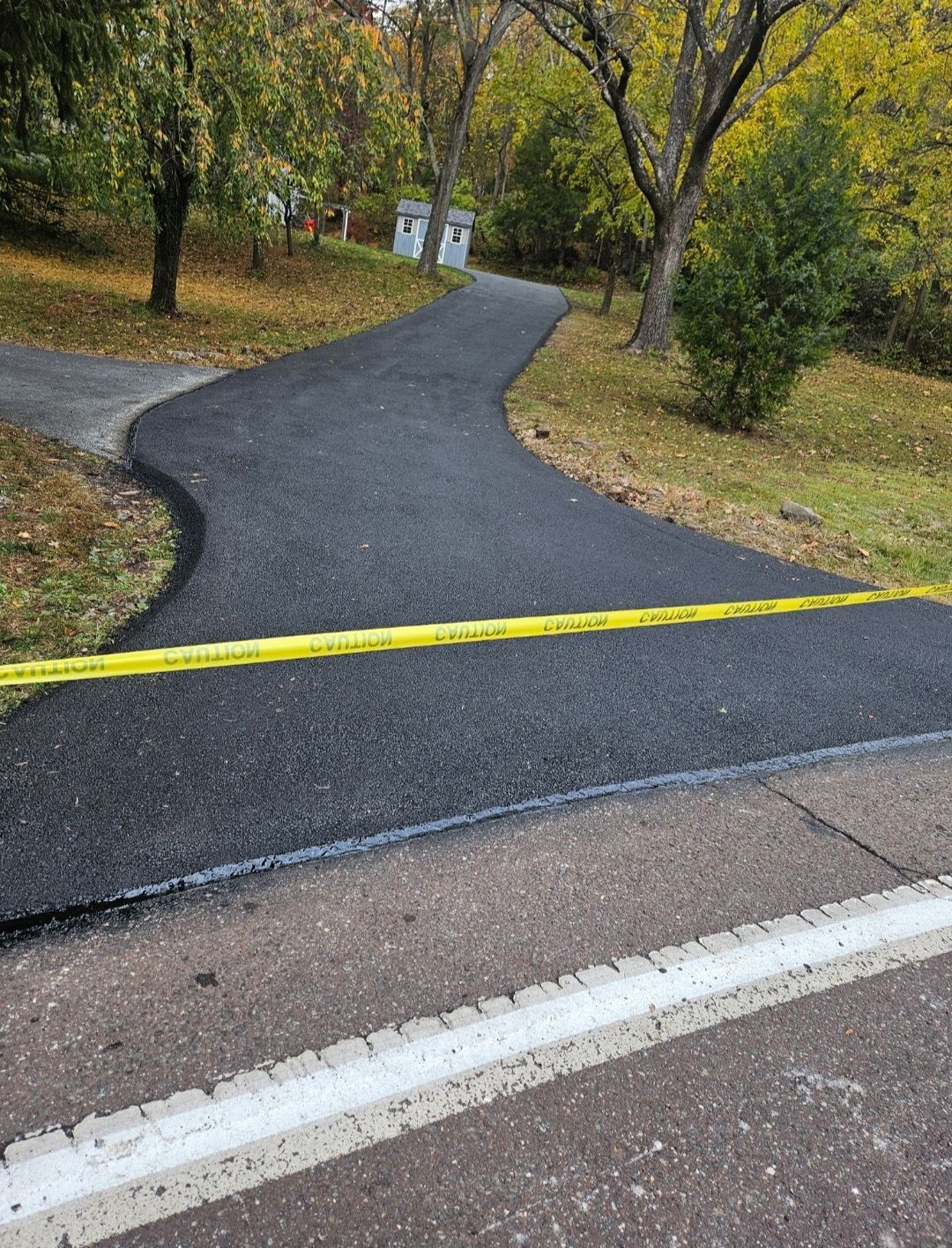 A freshly paved black asphalt path branches into two, blocked by a yellow caution tape, set against a wooded backdrop.