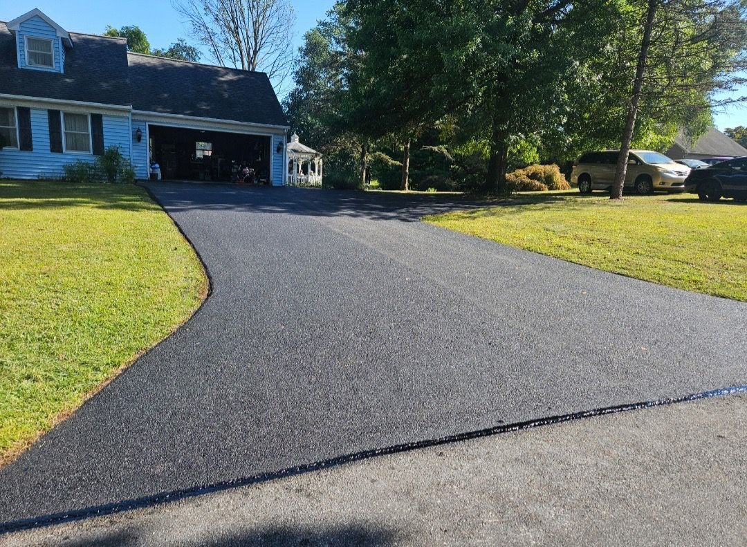 A freshly paved asphalt driveway leading to a blue house with a garage, flanked by green grass under a clear blue sky.