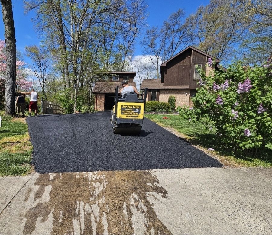 A yellow roller machine compacts fresh black asphalt on a residential driveway in front of a house on a sunny day.