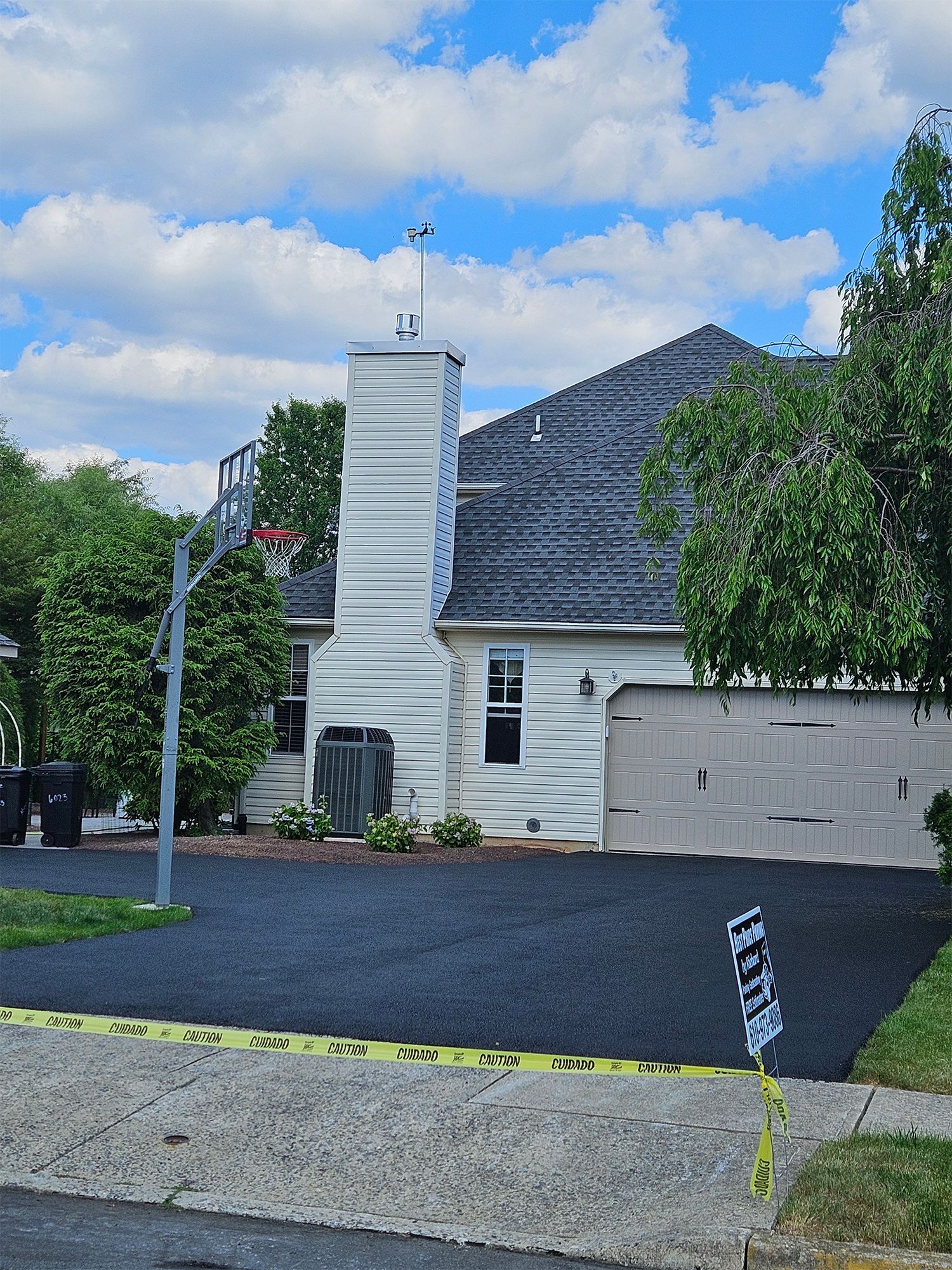 A freshly paved black driveway leads to a light-sided house with a large chimney and a basketball hoop.