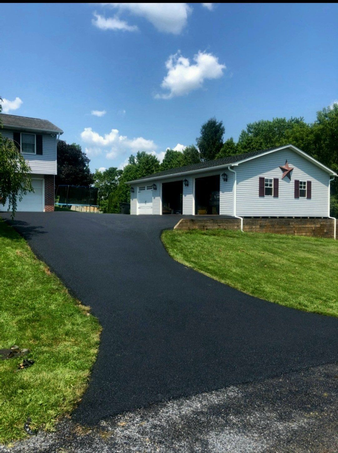 Black asphalt driveway leading to a light blue house and garage on a sunny day.