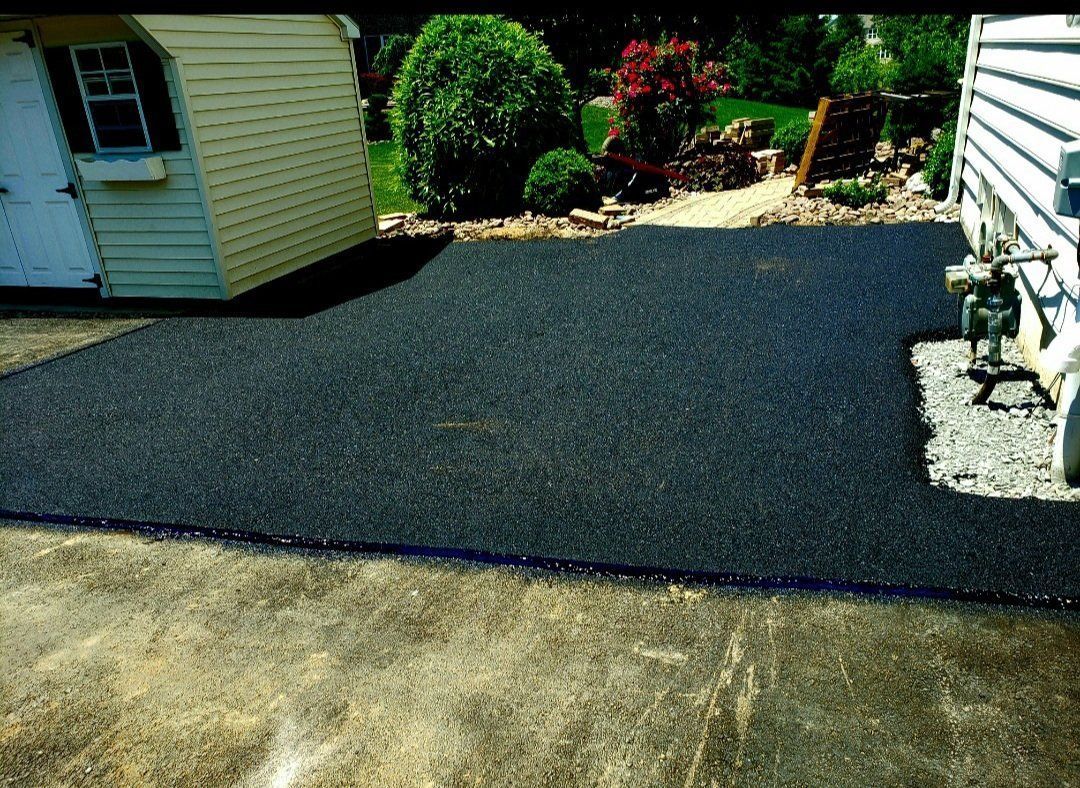 Black asphalt driveway, adjacent to a shed and a house. Green shrubbery in the background.