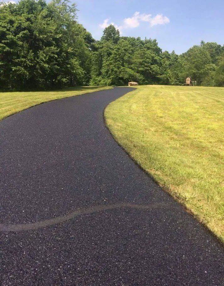 Black asphalt path winds through a grassy lawn, trees in the background under a blue sky.
