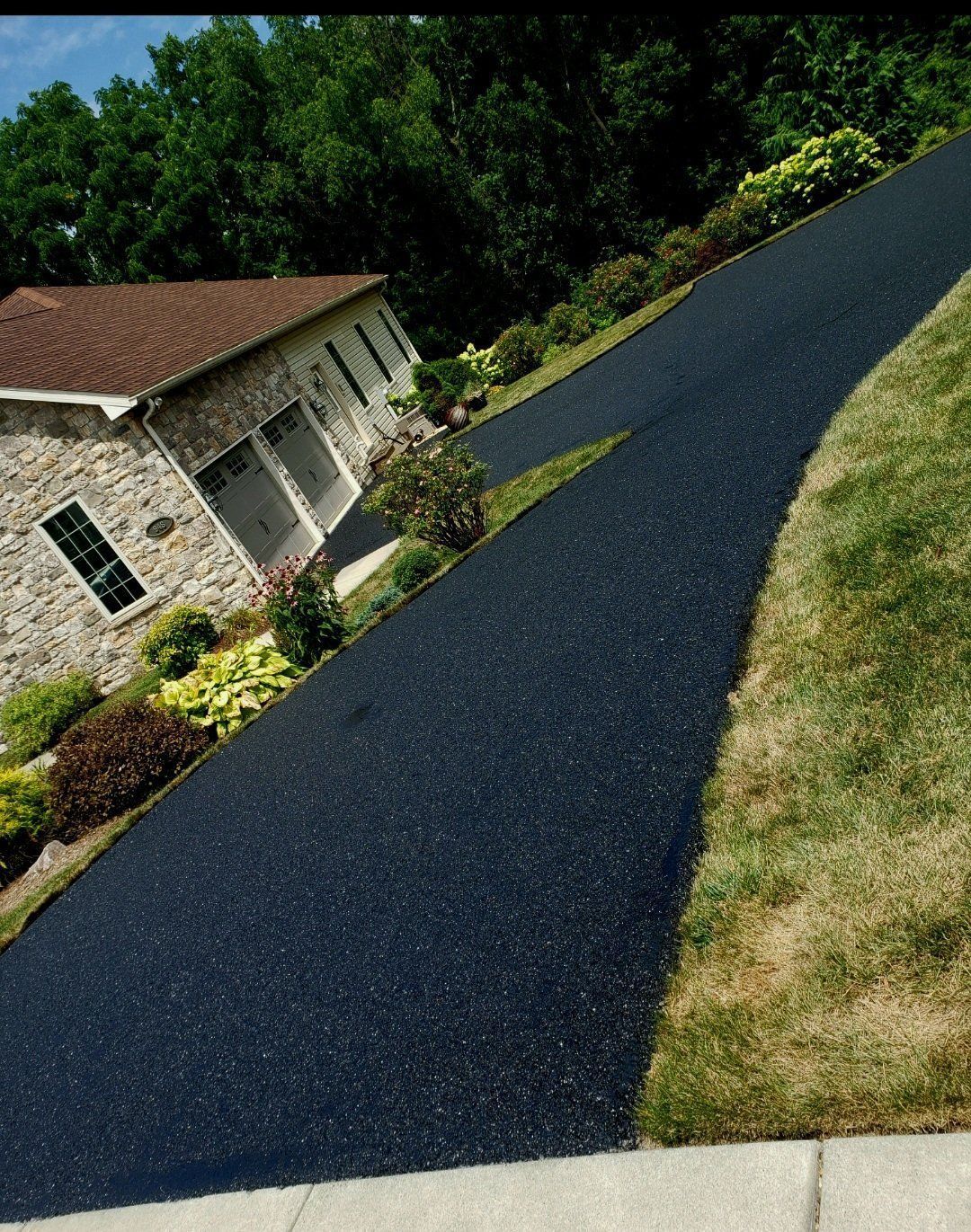 Black asphalt driveway leads up to a stone house with a garage, bordered by green grass and landscaping.