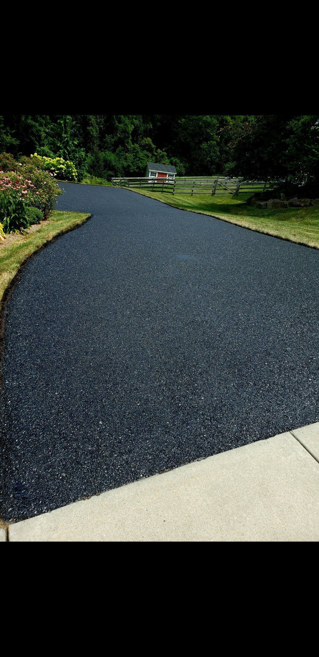 Black gravel driveway, bordered by grass, leads to distant trees. Concrete sidewalk in foreground.