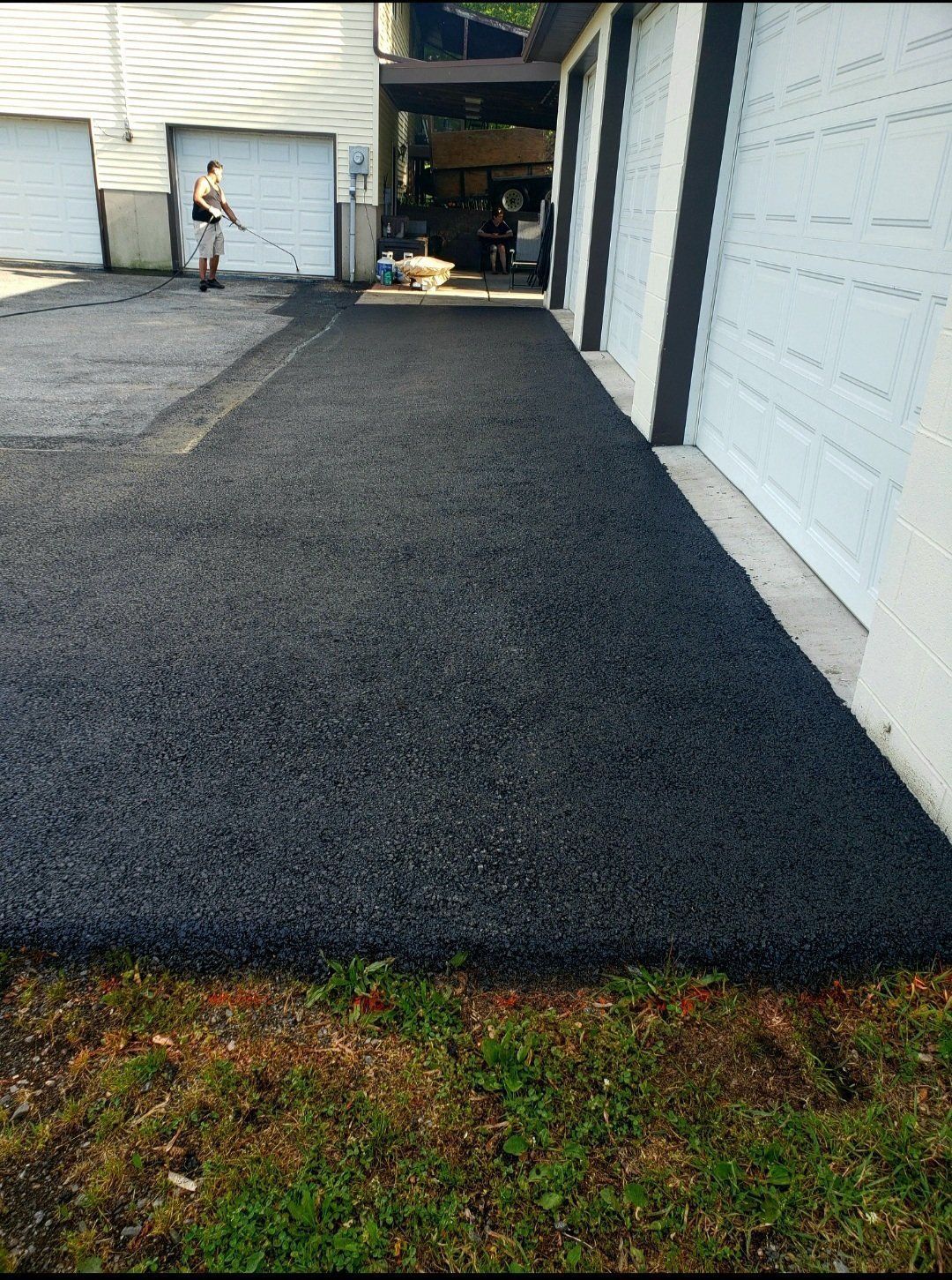 Freshly paved black asphalt driveway alongside a garage with white doors; a person sprays in the distance.