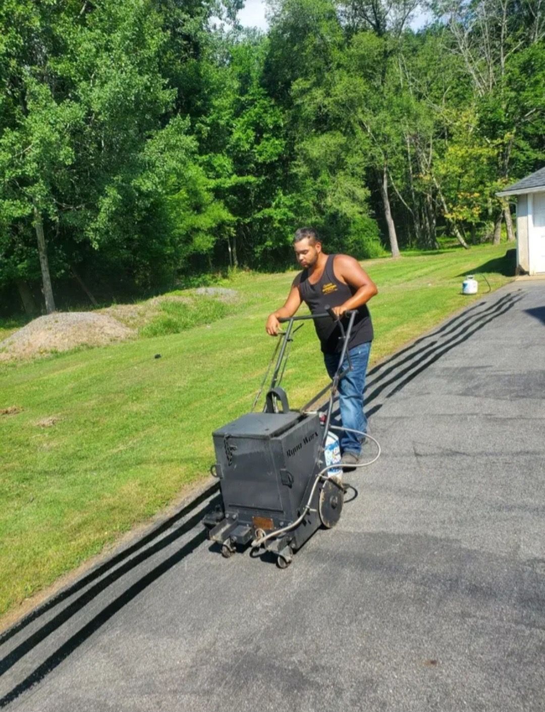 A person in a black tank top pushes a walk-behind asphalt crack sealer machine along a driveway next to a grass lawn.