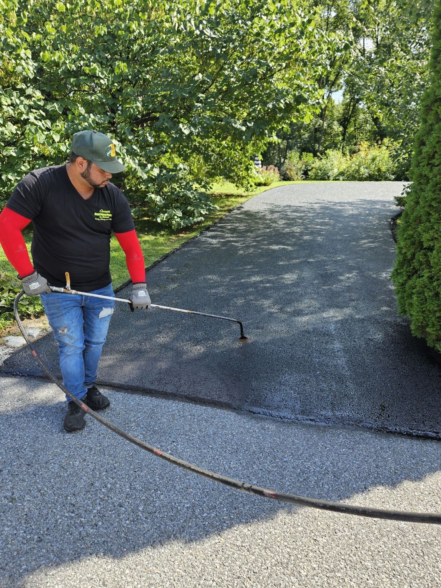 A worker in a cap and gloves uses a spray wand to apply sealant to a gravel driveway in a wooded residential area.