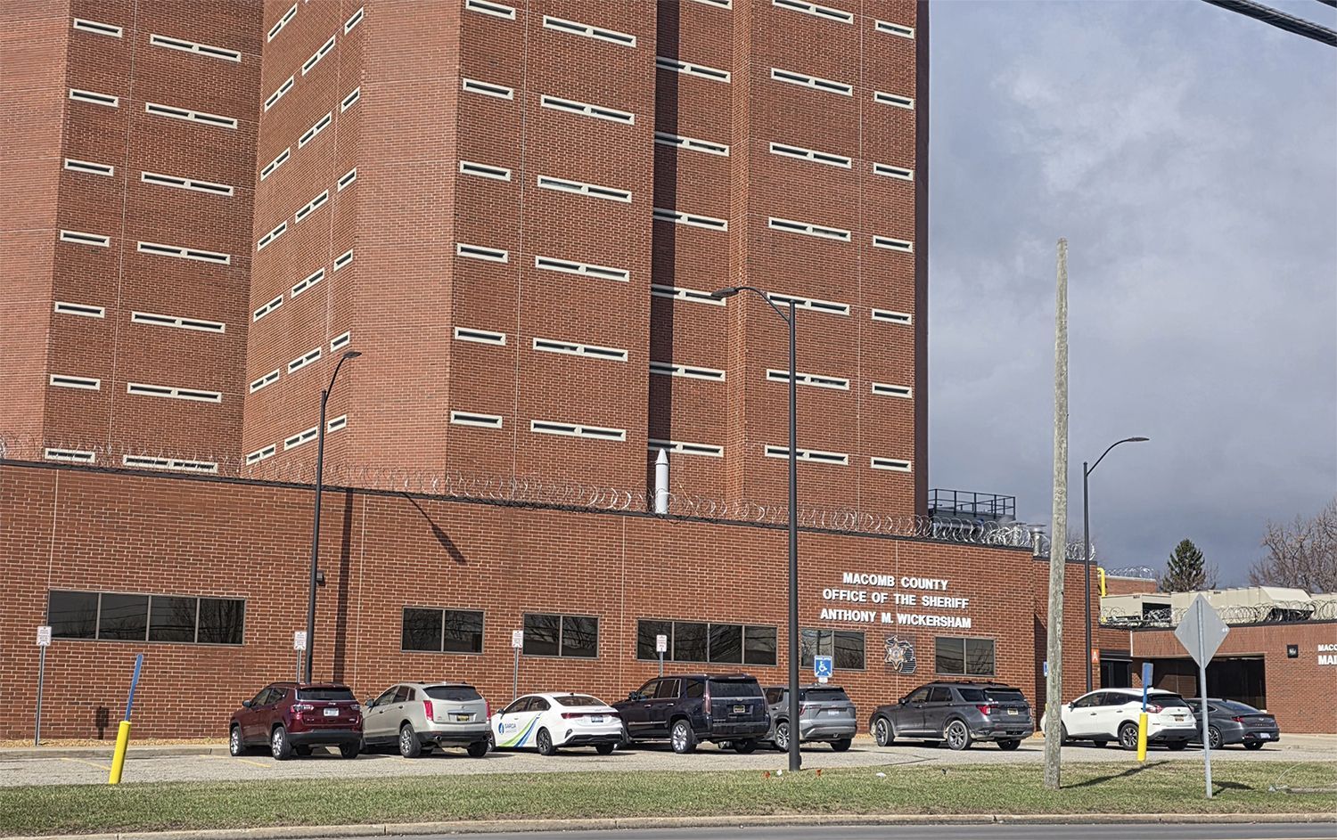 A multi-story brick building with rows of narrow windows, topped with barbed wire, and parked cars in the foreground.