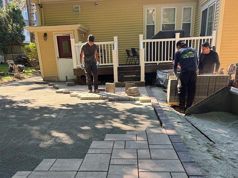 A Bobcat is cutting a tree branch that fell on a gray Audi parked on a brick-paved street.