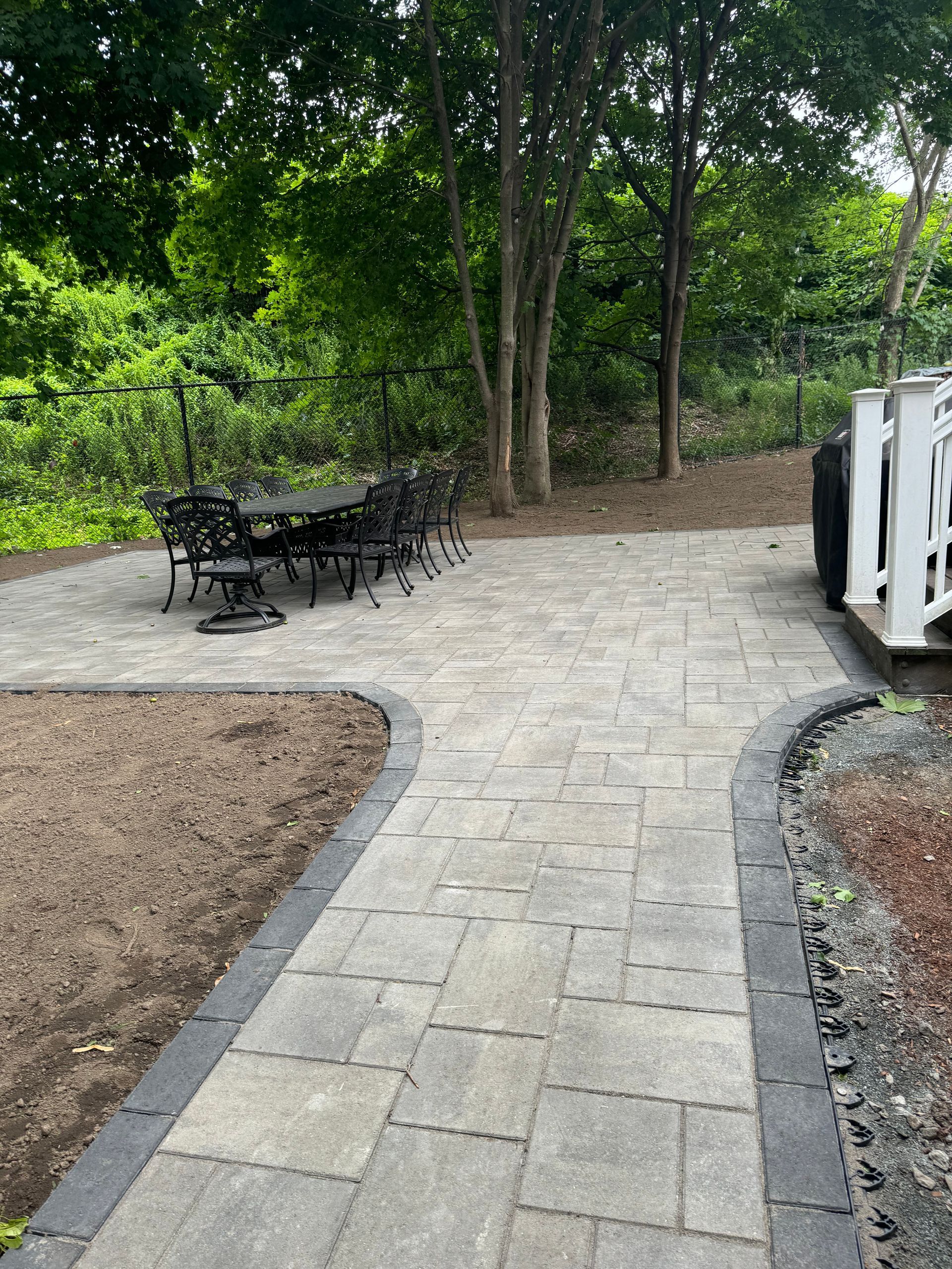 Stone patio with black table and chairs, path, trees, and white railing.