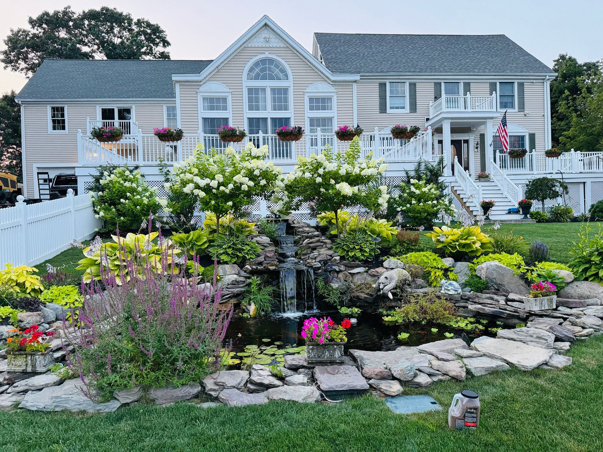 Two-story house with a white fence, waterfall garden, and hanging flower baskets.