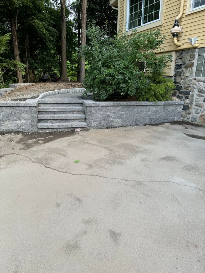 Concrete steps leading up to a retaining wall next to a house with a stone foundation and yellow siding.