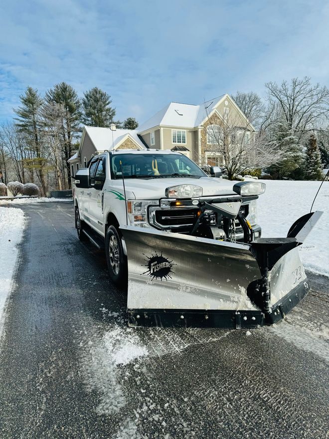 Person using a snow blower to clear a driveway in front of a snow-covered suburban house.