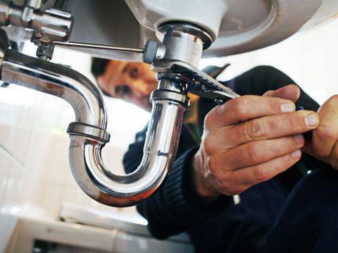 Plumber fixing a sink drain with a wrench. The chrome pipes and sink are visible.