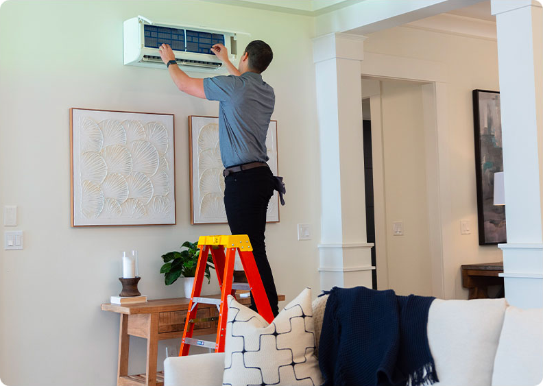 A technician on a ladder performs maintenance on a white wall-mounted air conditioning unit inside a living room.