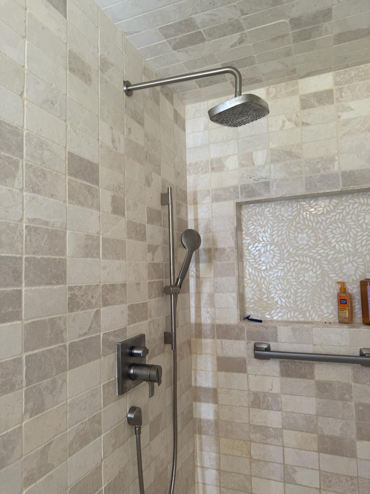 Shower with beige and gray tiled walls, rain shower head, handheld shower, recessed shelf.