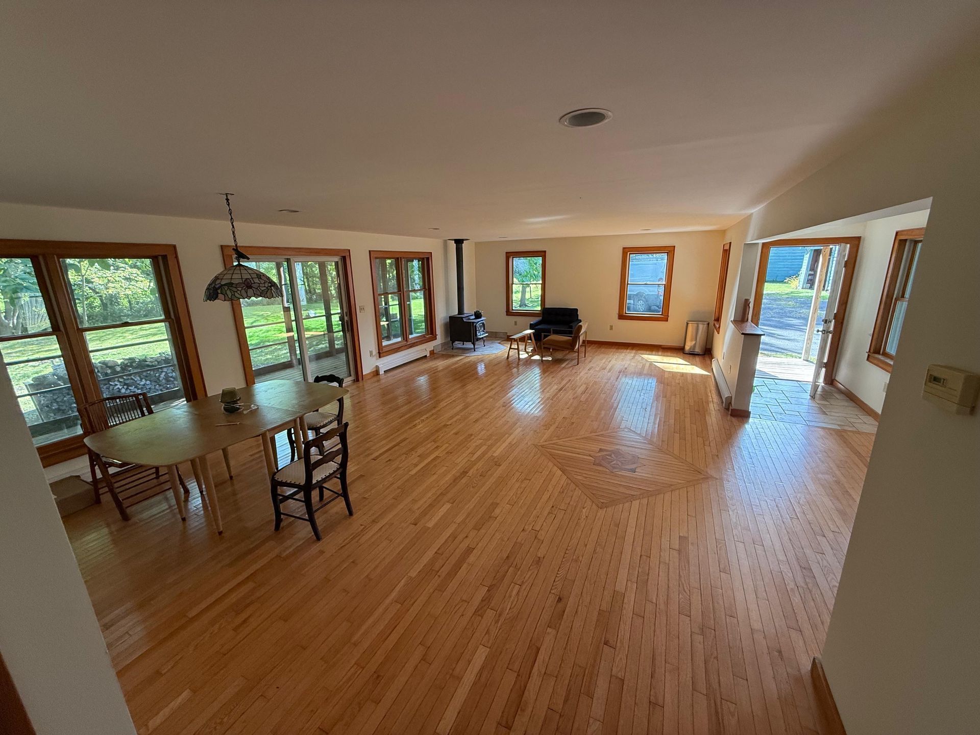 An open-concept room with light hardwood floors, a dining set, a wood stove, and large windows looking out to the outdoors.