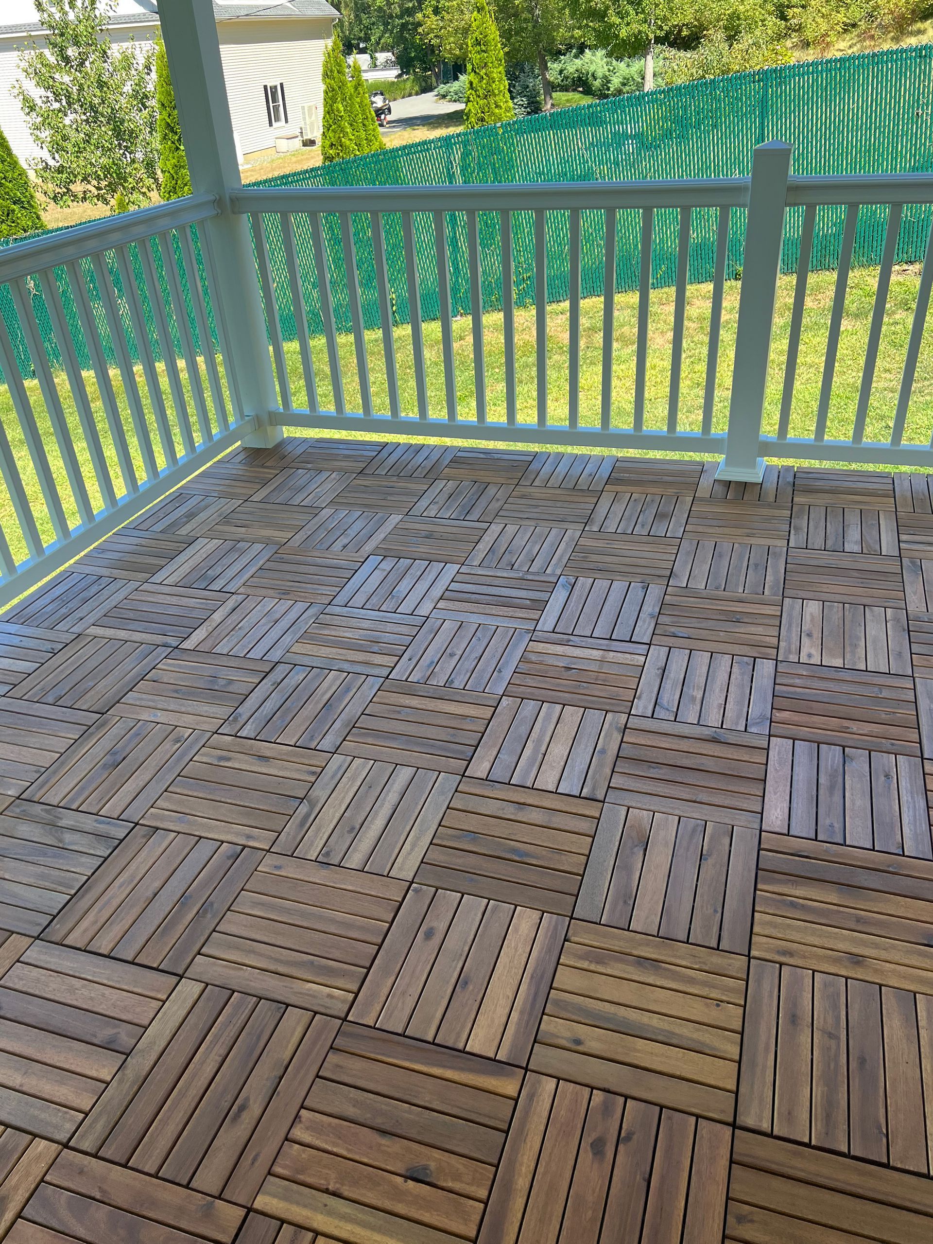 A wooden deck made of square interlocking tiles on a porch, with a white railing overlooking a green grassy yard.