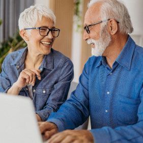 Smiling couple looking at laptop screen indoors. Woman wears glasses, denim shirt. Man wears glasses, blue shirt.