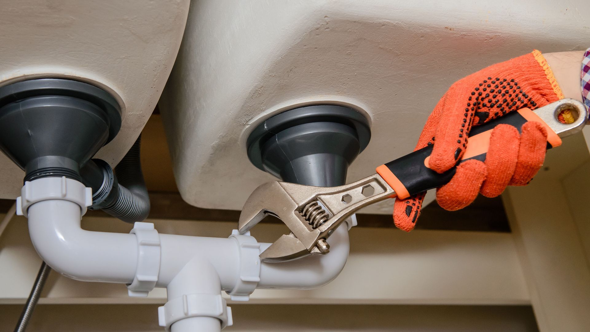 A person's gloved hand using a wrench to tighten a white PVC pipe under a kitchen sink.