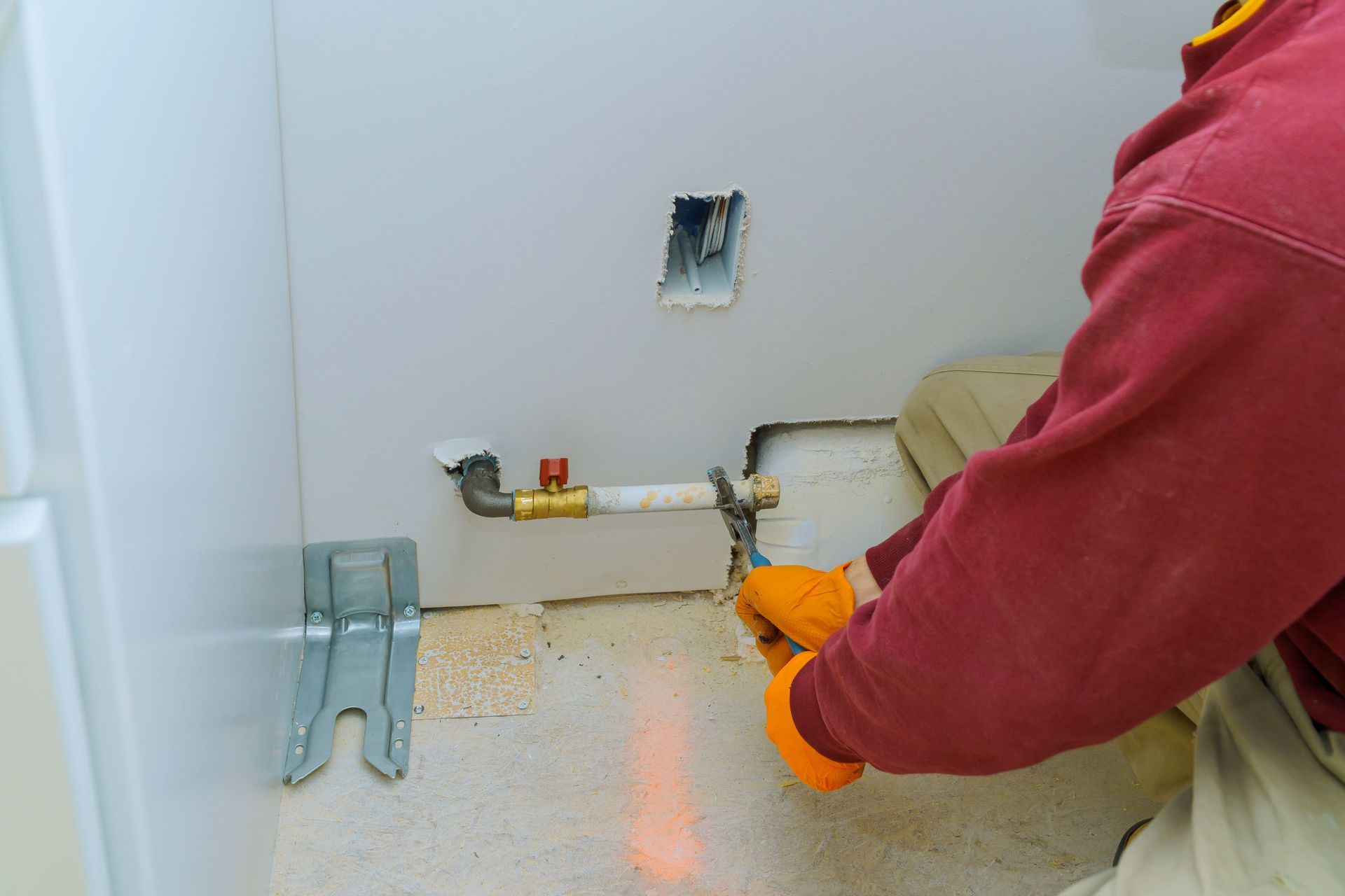 A person in orange gloves uses a wrench to tighten a pipe in a white-walled room, near an electrical outlet.
