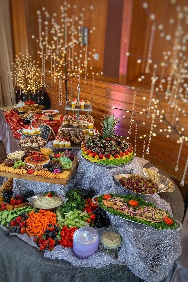 A multi-tiered catering display features assorted appetizers, fruit, and vegetables on a silver-draped table with fairy lights.
