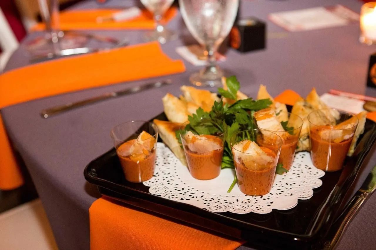 A black tray on a grey table holds small plastic cups of dip with crackers and a garnish of fresh green herbs.
