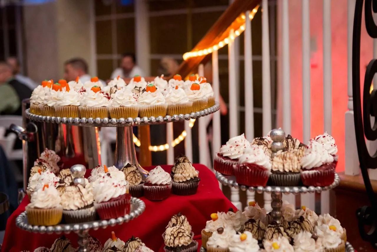 Tiered silver stands display an assortment of decorated cupcakes on a red-covered table at an indoor event.