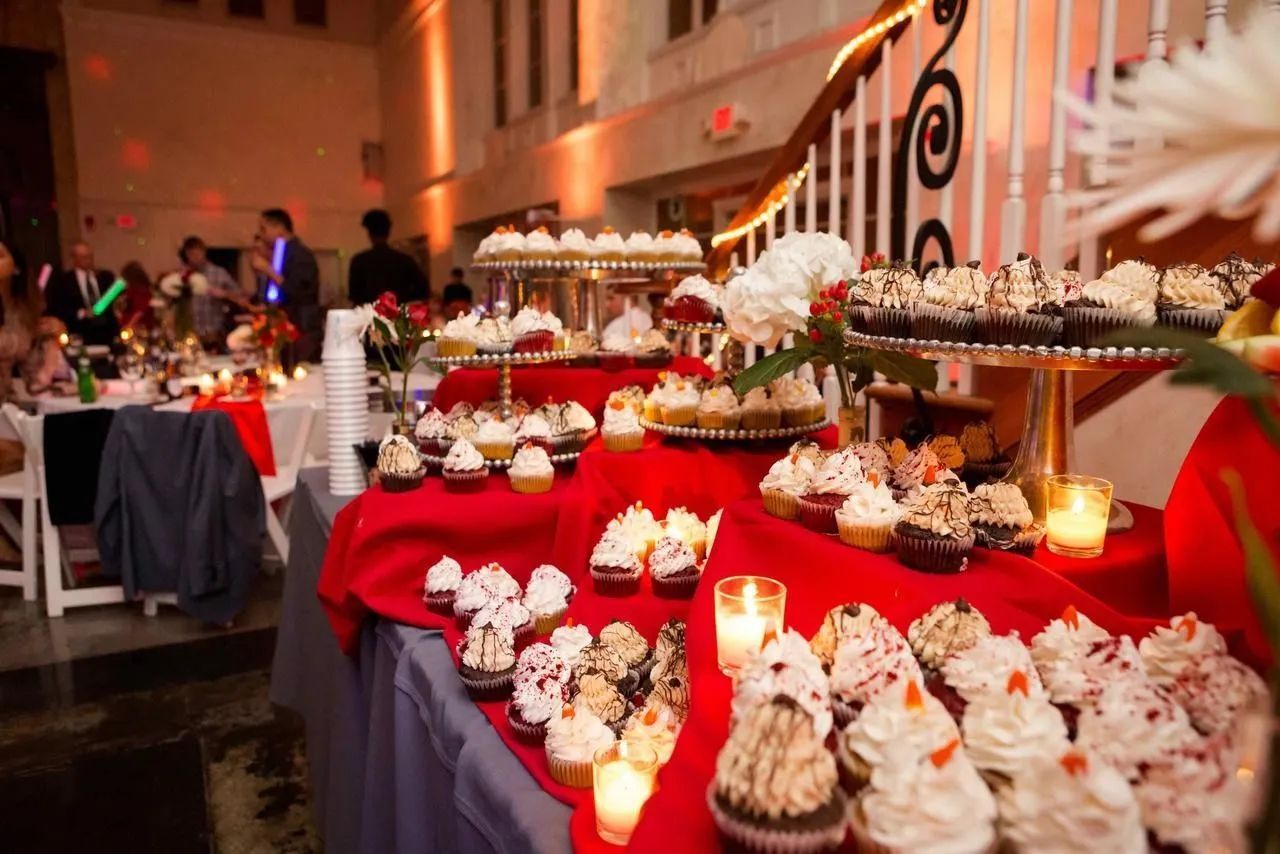 A buffet table with rows of cupcakes sits in a dimly lit, festive event space with blurred guests in the background.