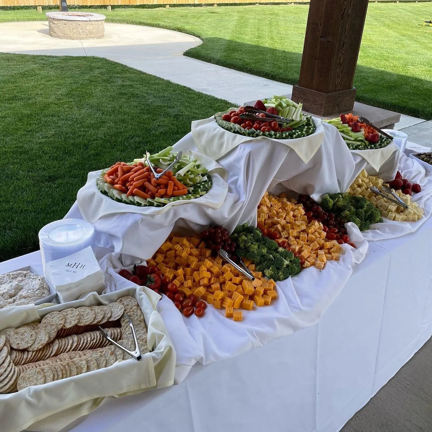 A multi-tiered outdoor buffet table featuring platters of cheese, crackers, and raw vegetables on a white tablecloth.
