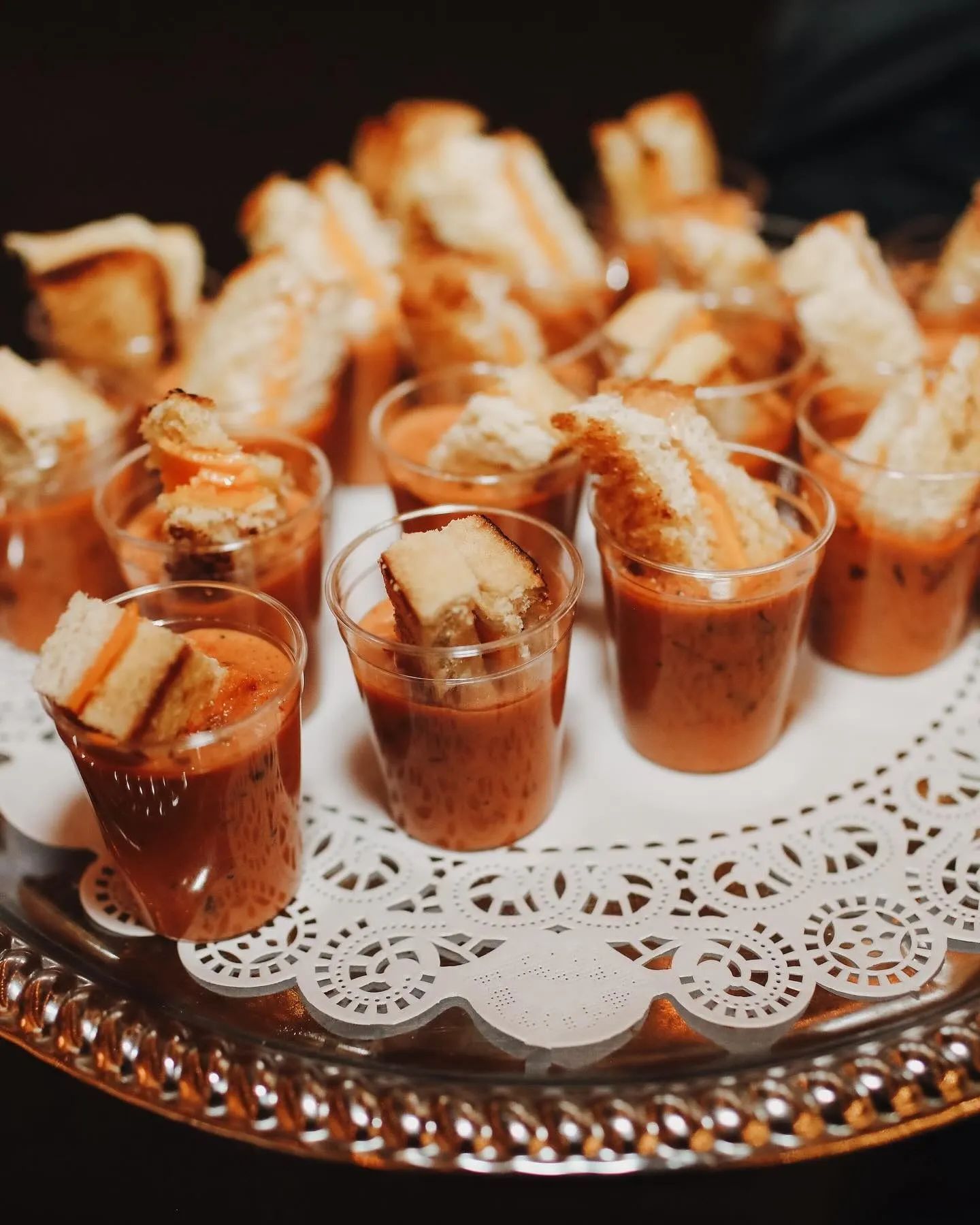 A silver platter holds several clear shot glasses filled with tomato soup, each topped with a small grilled cheese wedge.