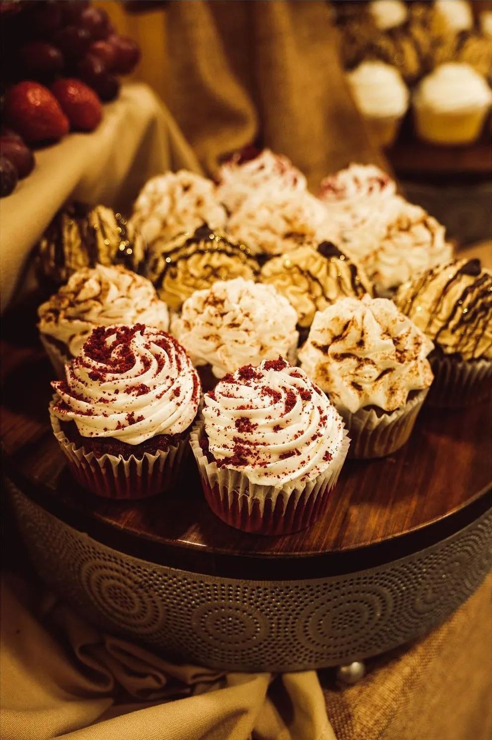 A tiered stand displays an assortment of decorated cupcakes with swirls of frosting, arranged on a rustic table.