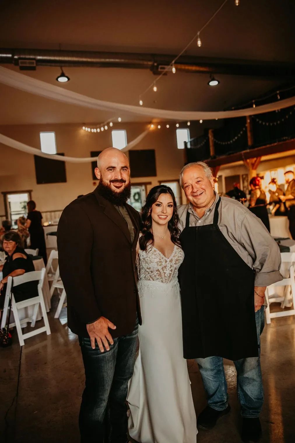 A man in a suit, a woman in a wedding dress, and a man wearing an apron stand together smiling in a festive event hall.