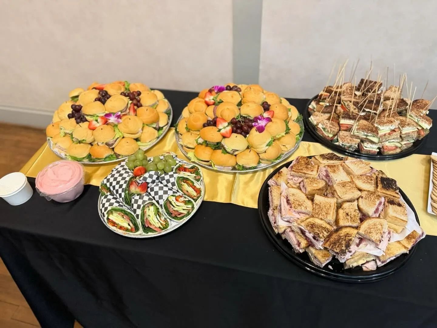 A catering table with several platters of sandwiches and wraps served on a black tablecloth with a yellow runner.