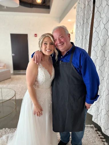 A smiling bride in a white lace wedding dress poses with an arm around a man wearing a blue shirt and a black apron.