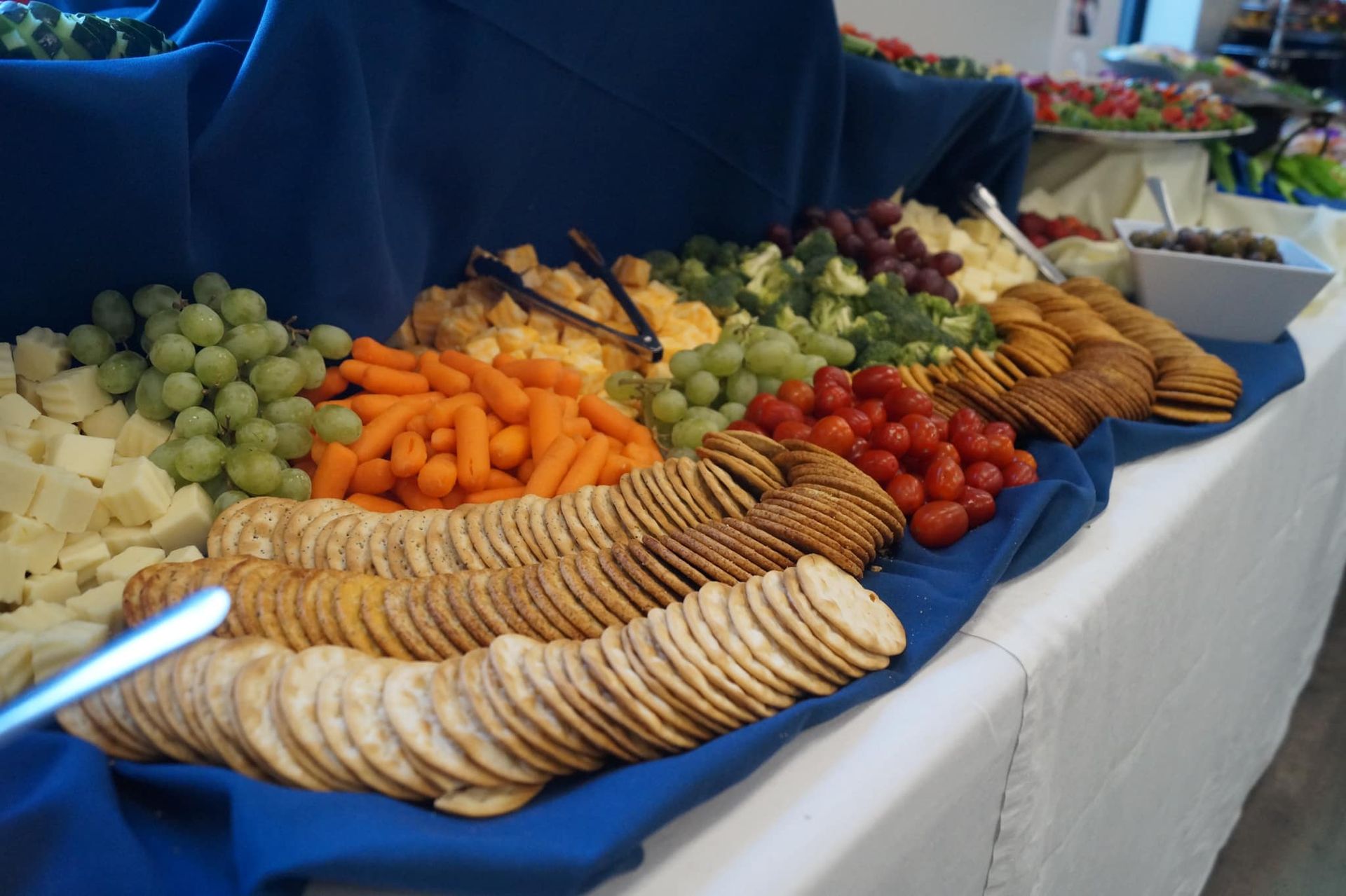 A buffet table with a blue tablecloth, featuring rows of crackers, cheese cubes, carrots, grapes, and cherry tomatoes.