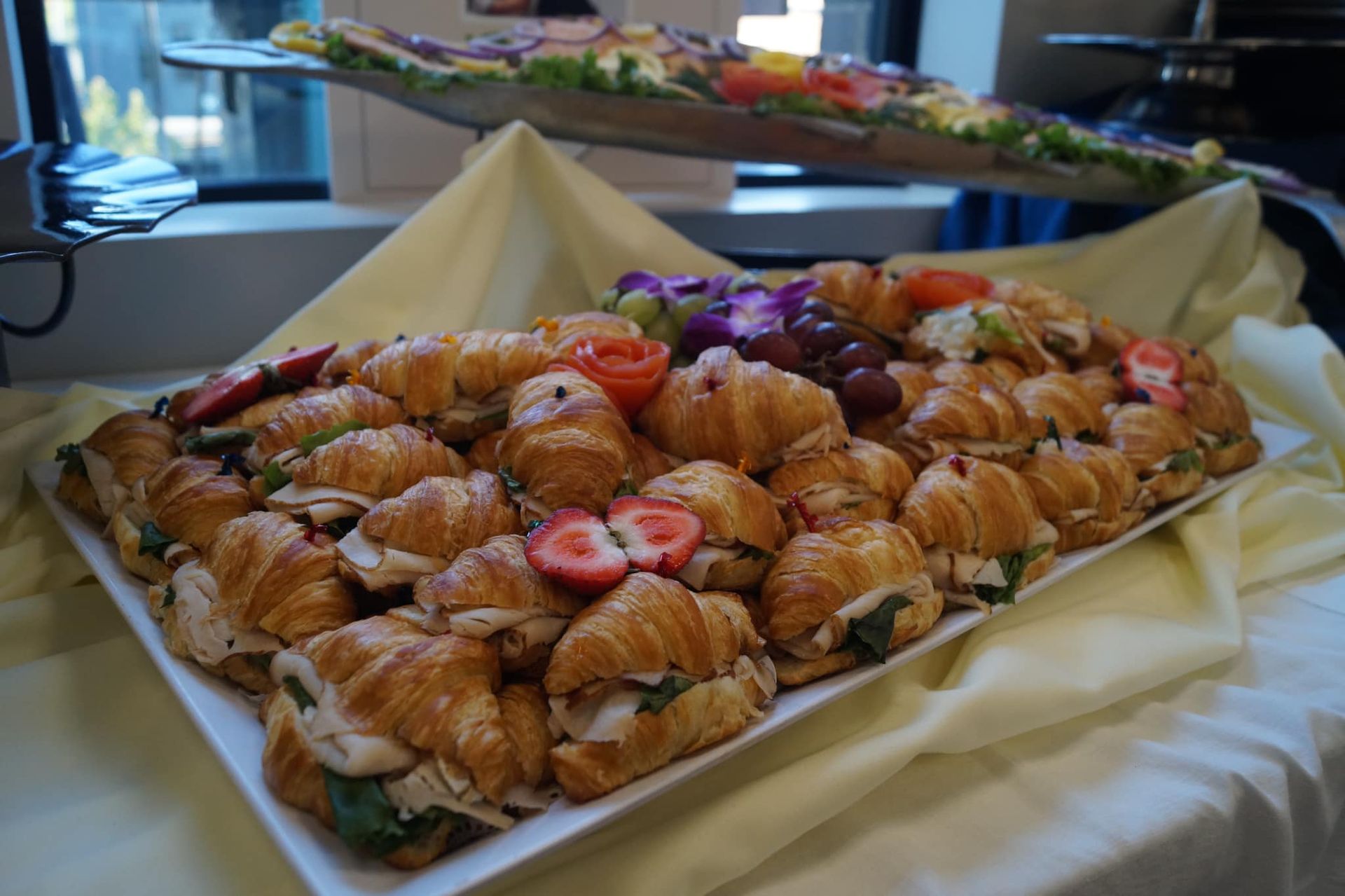 A tray of turkey croissant sandwiches garnished with strawberry slices and grapes, served on a light yellow tablecloth.