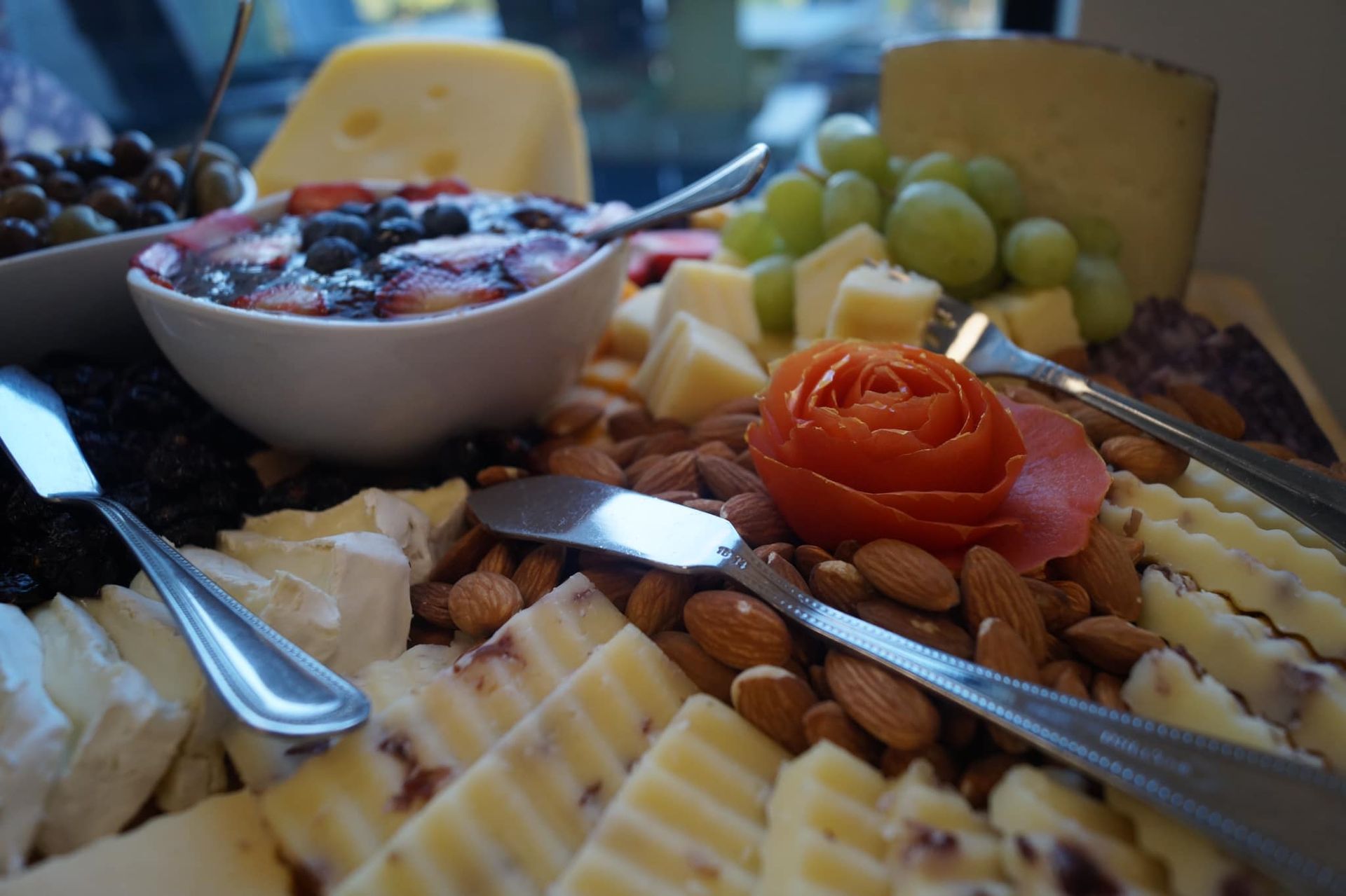 A charcuterie board featuring assorted sliced cheeses, green grapes, almonds, dark berries, and a dish of fruit compote.
