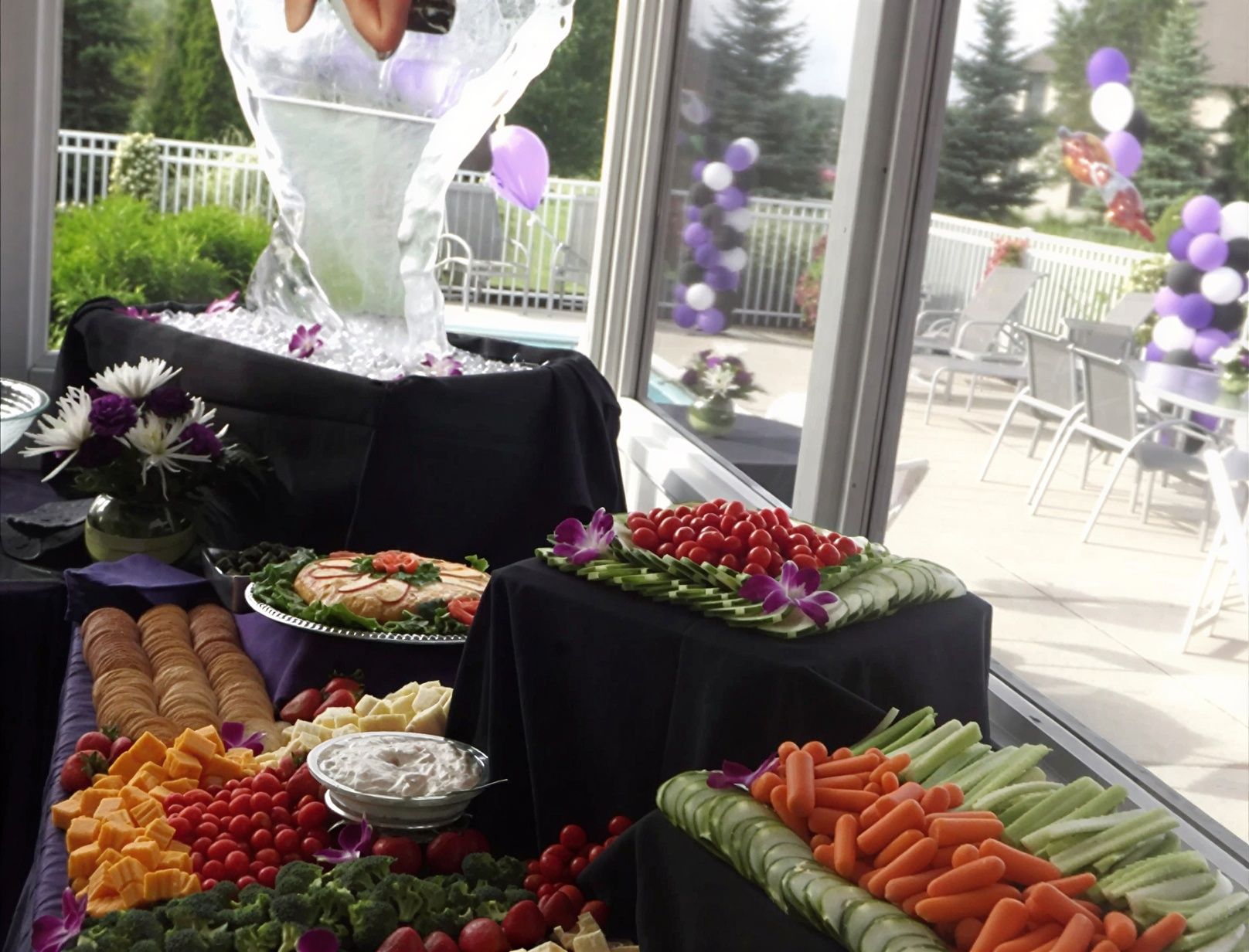 Elegant fruit and vegetable buffet on black tablecloths by a sunlit pool with purple balloons