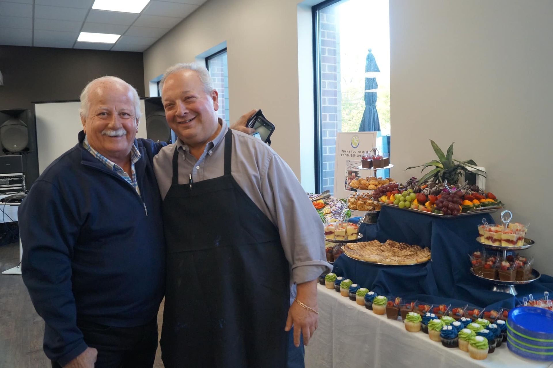Two smiling men stand together in front of a table filled with various appetizers and desserts in a bright indoor room.