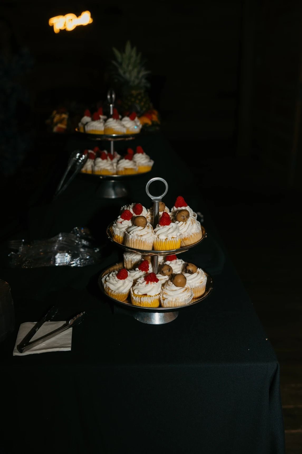 Tiered trays of cupcakes with white frosting and cherries on a black table in a dimly lit room