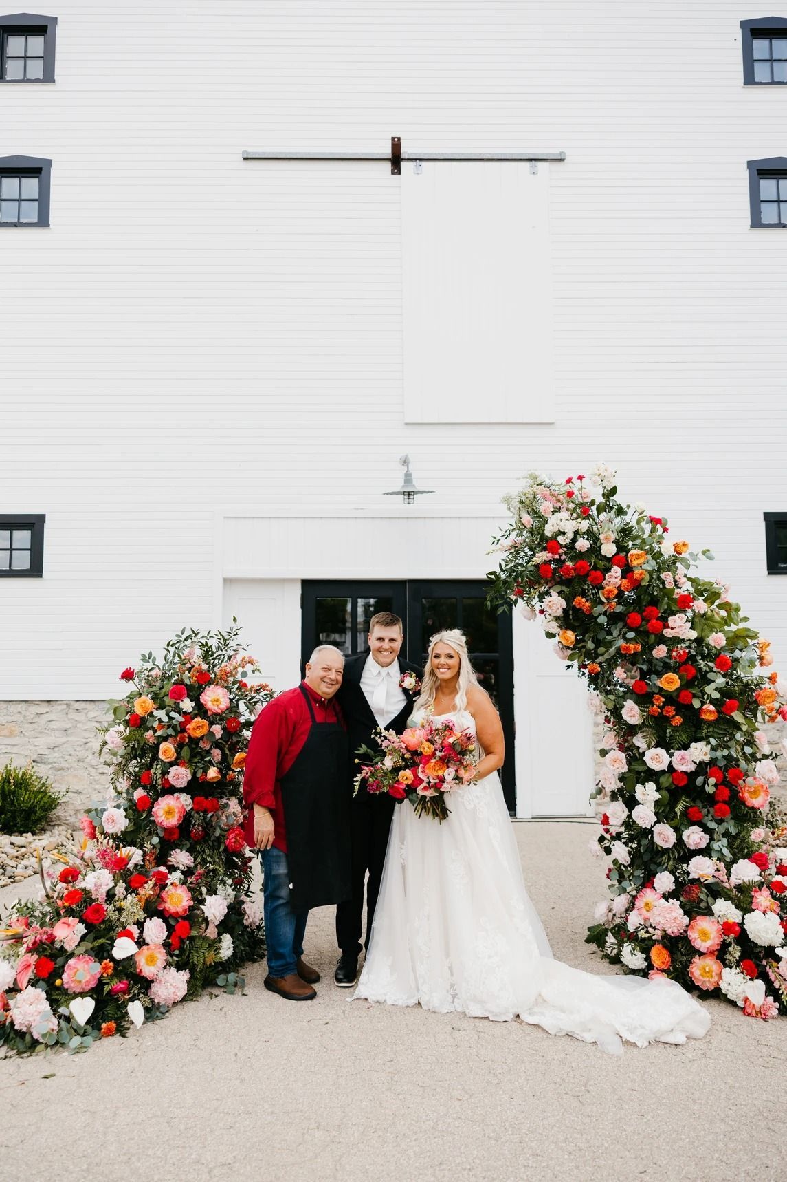 Wedding couple posing with an officiant under a floral arch at a white building entrance
