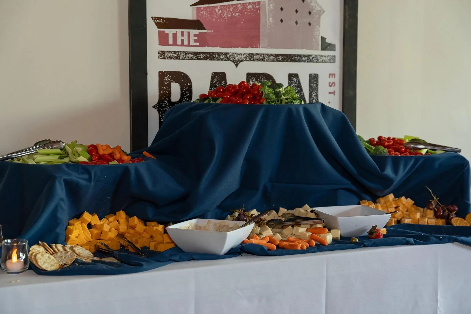 A buffet table with blue linens, featuring bowls of various cheeses, crackers, and vegetables set beneath a barn sign.