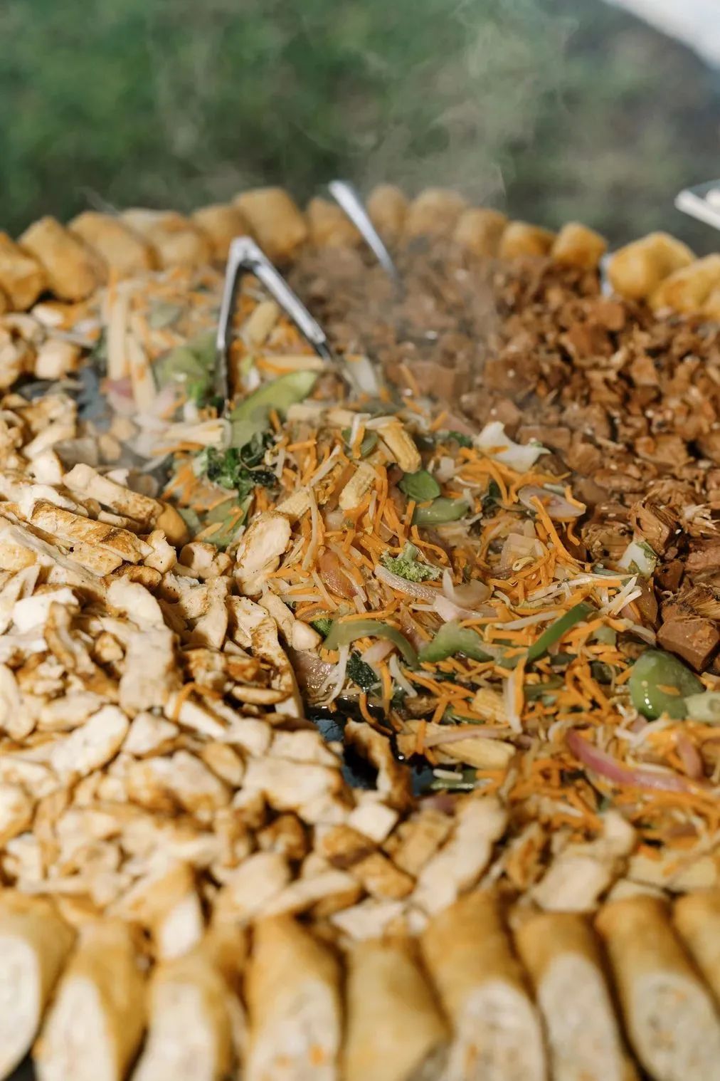 A circular grill filled with sliced meat, shredded carrots, vegetables, and spring rolls being tossed with metal tongs.