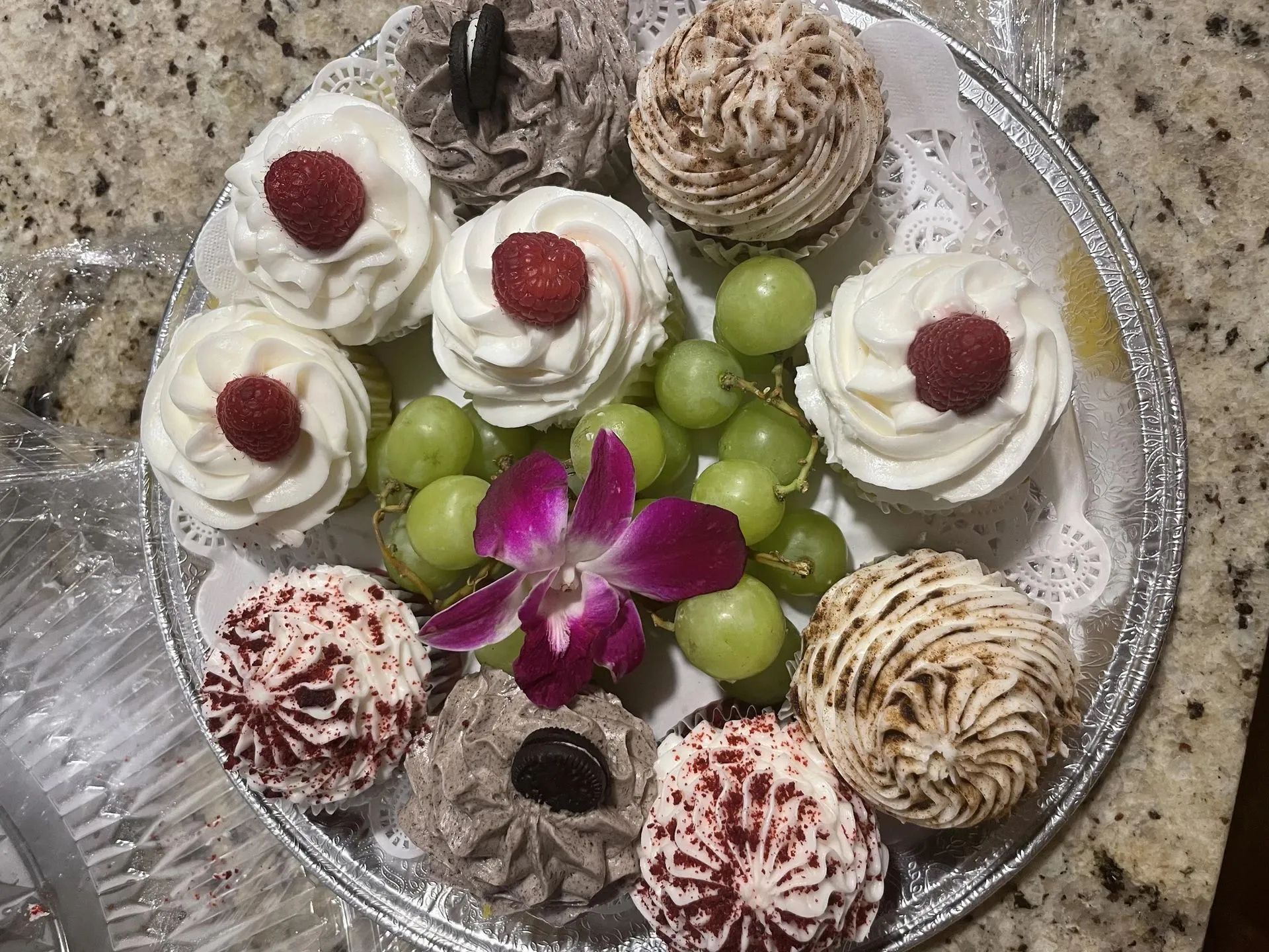 A platter of assorted cupcakes topped with berries and frosting, accented by a central purple orchid and green grapes.