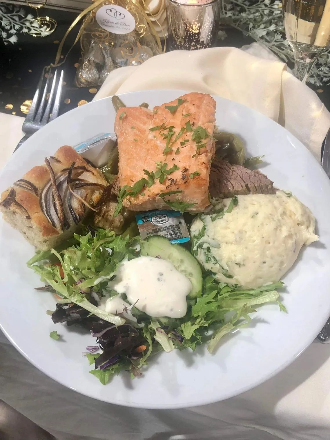 A plated meal featuring salmon, mashed potatoes, a small salad, and a slice of onion-topped focaccia bread.