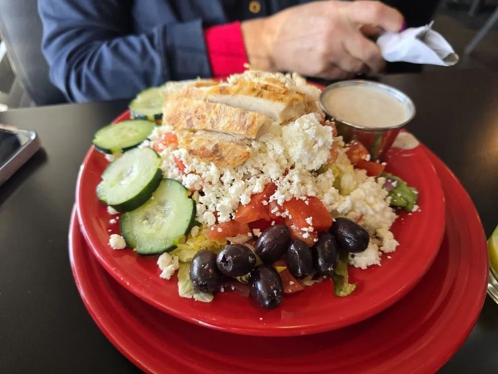 A Greek-style salad on a red plate, topped with sliced chicken, feta, olives, and cucumbers, with a side of dressing.